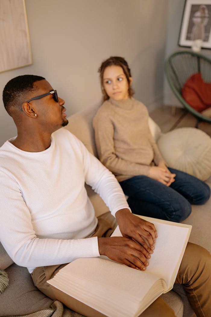 Man reading a Braille book while sitting on a sofa indoors, accompanied by a woman.