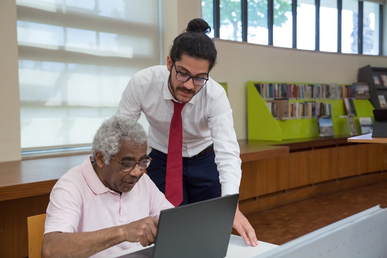 An elderly man learning to use a laptop with assistance in a library.